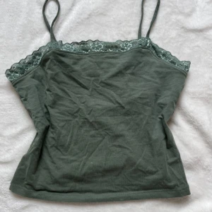 Olivgrön linnetopp med spets  - Säljer en olivgrön linnetopp från Kappahl med tunna axelband och dekorativ spets längs urringningen. Toppen är figurnära och i mjuk bomull, perfekt för varma dagar eller som lager under andra plagg.
