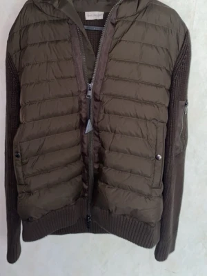 Olive grön jacka från moncler - Snygg grön jacka  från Moncler med stickade ärmar och quiltad front. Jackan har dragkedja, ribbade muddar och en ficka med dragkedja på ärmen. Perfekt för dig som vill ha en cool och varm look under höst och vinter. Priset går absolut att diskutera om snabb affär