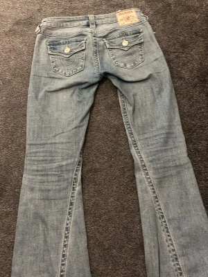 True Religion Joey ljusblå jeans - Ljusblå jeans från True Religion, modell Joey med bootcut-snitt. Snygga detaljer på bakfickorna med silverfärgade knappar och broderade sömmar. Klassisk femficksdesign och låg midja. Materialet är jeans med en tvättad look. Har ett litet hål på ena sidan annars väldigt bra skick❤️🥰