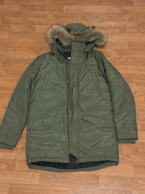 Grön parkajacka med fuskpäls - Säljer en grön parkajacka med huva och fuskpälsdetalj. Jackan har långa ärmar, stora fickor framtill och dragkedja. Perfekt för kalla dagar med sin varma och rejäla design. Stilen är klassisk och passar dig som gillar streetwear.