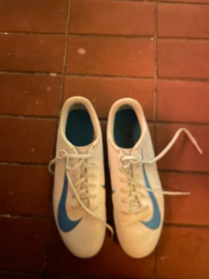 Nike fotbollsskor vit/blå strl 42.5 - Säljer ett par Nike fotbollsskor i vitt med blå detaljer och snörning. Skorna har en rund tå och platt sula med bra grepp, perfekt för spel på konstgräs eller inomhus. Materialet är syntet och skon har en klassisk Nike-logga på sidan och under sulan.