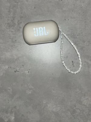 JBL trådlösa hörlurar med laddningsetui - Snygga och kompakta JBL true wireless-hörlurar i vitt med laddningsetui och handledsrem. Hörlurarna och etuiet är i fint skick utan synliga repor eller skador. Perfekt för musik, samtal och träning. Laddas via micro-USB.