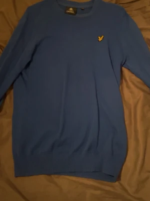 Blå stickad tröja från Lyle & Scott storlek S - Säljer en blå stickad tröja från Lyle & Scott med deras klassiska gula logotyp på bröstet. Tröjan har rund hals och långa ärmar, perfekt för dig som gillar stilrena och enkla plagg. Ribbstickad nederkant och ärmslut ger en snygg passform.