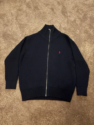 Blå stickad kofta från Polo Ralph Lauren - Säljer en blå, ribbstickad kofta från Polo Ralph Lauren i storlek M. Koftan har hel dragkedja framtill, hög krage och den klassiska röda Polo-loggan broderad på bröstet. Materialet är mjukt och stickat, perfekt för kyliga dagar.