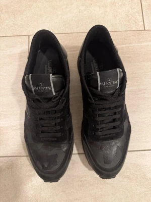 Valentino Garavani Rockrunner sneakers - Svarta Valentino Garavani Rockrunner sneakers med coola nitar på hälen och en snygg camouflagemönstrad ovandel. Skorna har snörning, rund tå och platt sula. Materialmix av mocka och skinn ger en lyxig känsla och street vibe.
