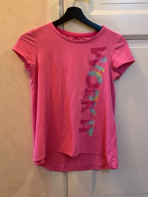 Rosa tränings-tshirt från Dobsom - Säljer en rosa t-shirt från Dobsom med trycket 'WORK IT' i färgglada bokstäver längs sidan. T-shirten har korta ärmar, rund hals och är tillverkad i ett lätt och skönt syntetmaterial som passar perfekt för träning. Modellen är något längre baktill för extra komfort.