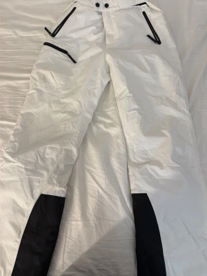 Vita täckbyxor från Cross Sportswear - Säljer ett par vita täckbyxor från Cross Sportswear i storlek M. Byxorna har svarta detaljer, dragkedjor och förstärkningar vid bensluten. Utrustade med svarta hängslen med logga och flera praktiska fickor med dragkedja. Perfekta för vinterns äventyr. Tyvärr har byxorna lite fläckar som syns på bild, men går enkelt bort vid tvätt