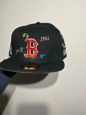 Svart Boston keps New Era - Säljer en svart Boston Red Sox keps från New Era med broderade detaljer som stjärnor, text och en eldslåga. Kepsen har röd B-logga framtill, broderad BOS-text på sidan och MLB-logga bak. Flat skärm och cool streetstil, perfekt för dig som gillar sport och snygga accessoarer.
