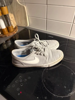 Gråa Nike sneakers med vit swoosh - Säljer ett par gråa Nike sneakers med klassisk vit swoosh på sidan. Skorna har snörning och är låga i modellen. Ovandelen är i textil och insidan har en blå detalj. Perfekta för dig som gillar stilrena och enkla sneakers.
