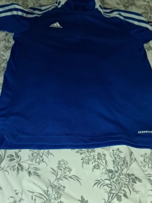 Blå Adidas Aeroready träningströja - Säljer en blå Adidas Aeroready träningströja med vita ränder på axlarna och Adidas-logga på bröstet. Tröjan har half zip och är gjord i ett syntetmaterial som andas. Perfekt för sport och träning, med klassisk Adidas-design.