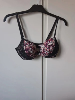 Blommig bh med svarta spetsdetaljer - Snygg bh med blommigt mönster i rosa, vit och svart. Kuporna har svarta spetsdetaljer på sidorna och justerbara axelband. Baksidan har klassisk knäppning och en liten öppning mellan kuporna. Perfekt för dig som gillar färg och mönster.