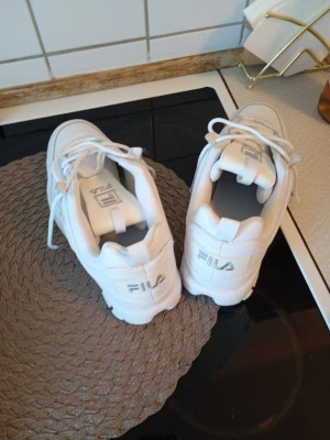 Vita chunky sneakers från Fila - Säljer ett par vita chunky Fila sneakers med platt sula och rund tå. Skorna har snörning och är tillverkade i syntetmaterial med Fila-logga på både plös och häl. Perfekta för dig som gillar en sportig och trendig stil.