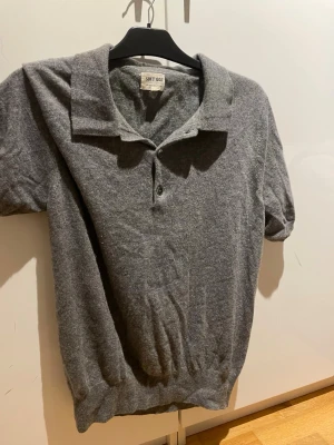 Grå kashmirpiké från Soft Goat - Mjuk och stilren grå pikétröja från Soft Goat i 100% kashmir. Tröjan har korta ärmar, klassisk krage och knäppning med två knappar framtill. Perfekt för dig som gillar en clean look med lyxig känsla.