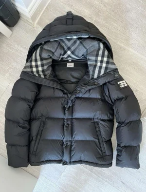 Svart dunjacka från Burberry - Snygg svart dunjacka från Burberry med klassiskt rutigt foder i huvan. Jackan har dragkedja och knappar framtill, två fickor med dragkedja och en mysig huva. Perfekt för kalla dagar och riktigt stilren look.