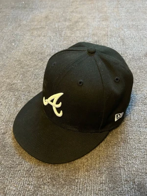 Atlanta Braves New Era 59FIFTY keps - Säljer en Atlanta Braves New Era 59FIFTY keps med ett broderat vitt 'A' framtill och MLB-logga baktill. Kepsen är tillverkad i 100% polyester och har en klassisk, rak skärm. Perfekt för dig som gillar streetwear och sportig stil.
