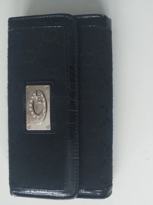 Svart plånbok med glansig finish - Snygg svart plånbok från Guess med glansig krokopräglad yta och detaljer i metall framtill. Plånboken har flera fack för kort, sedlar och mynt, samt genomskinliga fönster för ID. Stängs med tryckknapp och dragkedja.