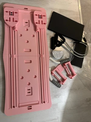 Rosa träningsbräda med handtag - Säljer en rosa träningsbräda med måtten 105x40 cm, perfekt för hemmaträning. Brädan har grå handtag och motståndsband, samt mjuka fotplattor. Den är hopfällbar och lätt att förvara under soffan. Materialet är slitstark syntetplast och handtagen har skumgrepp för extra komfort. Pilatesbrädan är helt ny men har en liten spricka, detta påverkar dock inte användningen
