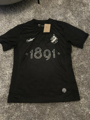 Svart Aik fotbollströja  - AIK x Nike fotbollströja i svart med tryck '1891' och klubbmärke på bröstet. Dri-FIT material som andas, kort ärm och rund hals. Snygg detalj med 'SMOKINGLIRARE' på ryggen. Perfekt för dig som gillar stilrena sportplagg.