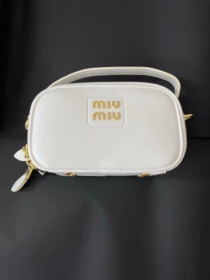 Vit handväska från Miu Miu - Säljer en stilren vit handväska från Miu Miu med guldiga detaljer och dubbla dragkedjor. Väskan är rektangulär med ett handtag upptill och har logotypen i guld på framsidan. Tillverkad i skinn med metallfötter undertill för extra lyxig känsla.