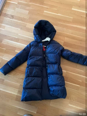 Mörkblå dunjacka från Polo Ralph Lauren - Säljer en lång, mörkblå dunjacka från Polo Ralph Lauren med stor huva och dragkedja framtill. Jackan har stora, quiltade partier och ett coolt rött POLO-tryck på insidan. Perfekt för kalla dagar när du vill hålla dig varm och se snygg ut.