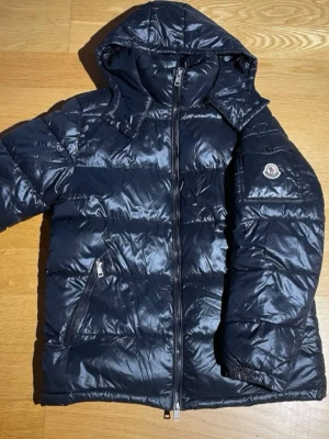 Svart Moncler pufferjacka med huva - Säljer en svart pufferjacka från Moncler med glansig finish och huva. Jackan har två sidofickor med dragkedja, Moncler-logga på ärmen och quiltad design. Fylld med vitt gåsdun för extra värme, perfekt för kalla dagar. Stängs med dragkedja framtill. Jackan är utrustad med NFC-chip i armmärket samt QR-kod på orginaletiketterna för verifierbar äkthet. Pris kan diskuteras vid smidig och snabb affär.