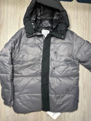 Grå Moncler pufferjacka med huva - Säljer en grå pufferjacka från Moncler med stor huva och svarta detaljer. Jackan har quiltad design, tryckknappar och en bred svart panel med Moncler-logga framtill. Materialet är glansigt och känns riktigt lyxigt, perfekt för kalla dagar.