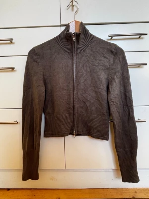 Grön stickad kofta från Junkyard - Säljer en militärgrön stickad kofta från Junkyard i storlek XS. Koftan har ribbad hög krage, långa ärmar och breda ribbade muddar vid ärmslut och nederkant. Dragkedja framtill som går hela vägen upp till kragen. Viskos/ polyamidblandning. Storlek XS. 