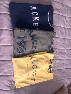 Tre Hackett tröjor med tryck - Säljer tre snygga tröjor från Hackett i storlek 13-14 år. En gul med marinblå logga, en olivgrön med broderat årtal och en mörkblå med vit text och röd/vit detalj vid kragen. Alla har rund hals och är kortärmade, perfekta för dig som gillar stilrena och coola prints. Köp antingen 1 för sig eller 3 för 600 totalt