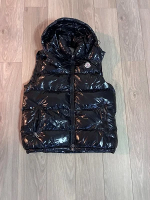 Moncler dunväst med luva + kvitto - Säljer en glansig svart dunväst från Moncler i storlek M. Västen har en stor, mysig huva, två fickor med dragkedja och ett klassiskt Moncler-märke på bröstet. Materialet är polyester och dun, perfekt för kyliga dagar. Stängs med dragkedja framtill.