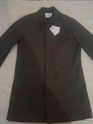 Svart rock från Bläck, storlek 48 Medium  - Stilren från Bläck med klassisk krage och diskreta sidofickor. Jackan har en rak passform och är tillverkad i ett slätt syntetmaterial som ger en clean look. Perfekt för dig som gillar minimalistisk stil och vill ha något som funkar till många outfits. olivgrön/mörkgrön storlek 48 helt ny Butikpris 1999kr