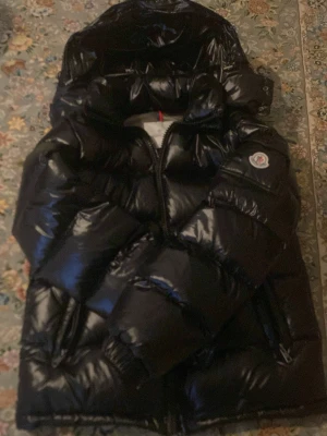 Moncler Maya  - Säljer en svart, glansig pufferjacka från Moncler med huva och dragkedja. Jackan har en klassisk Moncler-logga på ärmen och är fylld med dun för extra värme. Perfekt för kalla dagar och har en riktigt snygg streetstil.