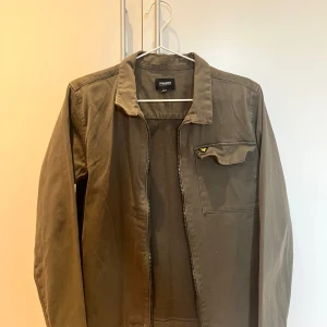 Olivgrön overshirt från Lyle & Scott - Säljer en olivgrön overshirt från Lyle & Scott Junior med klassisk krage och dragkedja framtill. Jackan har en bröstficka med lock och den ikoniska Lyle & Scott-loggan i gult. Materialet är mjuk bomull och modellen är normal i passningen. Hör av er vid eventuella frågor. Priset kommer inte ändras och är fast vid 200kr.
