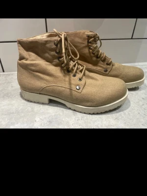 Vagabond 37 - Säljer ett par beige boots med snörning och rund tå. Skorna har en platt sula i ljus färg och är tillverkade i ett slitstarkt bomullsmaterial. Metallöglor och klassisk boot-design gör dem perfekta för dig som gillar enkel och clean stil.