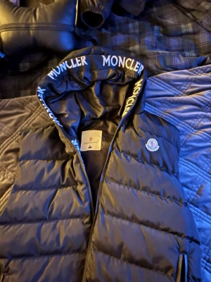 Svart dunväst från Moncler med huva - Säljer en svart dunväst från Moncler i storlek L. Västen har en vadderad, quiltad design, huva med Moncler-logga runt kanten och dragkedja framtill. Insidan har en röd dragkedjeficka och ett serietidningsmärke. Perfekt för dig som vill ha en stilren