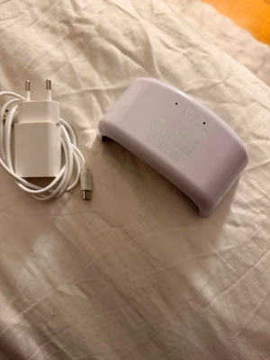 Depend Gel iQ UV/LED lampa - Säljer en vit Depend Gel iQ UV/LED lampa för gelnaglar, perfekt för att härda gel-lack hemma. Kommer med tillhörande vit USB-laddare och adapter. Lampan har en smidig och kompakt design som gör den enkel att använda och förvara.