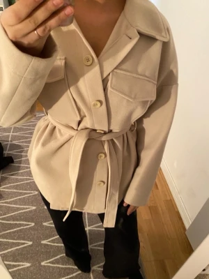 Beige overshirt med bälte och knappar - Snygg beige overshirt med stora bröstfickor, knappar framtill och matchande bälte i midjan. Jackan har en avslappnad passform och långa ärmar, perfekt att slänga på sig över en hoodie eller t-shirt. Enkel och stilren design som passar till det mesta.