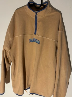Beige fleece-tröja ASOS - Säljer en beige fleece-tröja från ASOS DESIGN med blå detaljer och broderad Nashville-text fram och bak. Tröjan har half zip, lång ärm och blå kantband vid hals, dragkedja och nederkant. Perfekt för dig som gillar chill och streetwear.