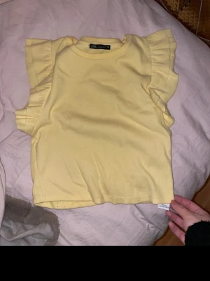 Gul volangtopp från Zara - Supersöt gul topp från Zara med volangdetaljer på ärmarna. Modellen är kortare och har en rund halsringning. Tillverkad i mjuk bomull som känns skön mot huden. Perfekt för dig som vill ha en färgklick i garderoben!