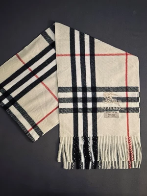 Rutig vit Burberry halsduk  - Säljer en klassisk Burberry halsduk i kashmir aktigt material med ikoniskt rutigt mönster i vitt, svart och rött. Halsduken har broderad Burberry-logga och fransar i ändarna. Perfekt accessoar för att lyfta din stil med lyxig känsla.