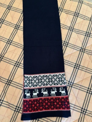 Mörkblå stickad halsduk från Malvin - Stickad halsduk från Malvin i mörkblått med klassiskt mönster i vitt och rött längst ner, inklusive renmotiv och geometriska former. Materialet är 70% polyakryl och 30% ull, vilket gör den både mjuk och varm. Perfekt för vintern och julens mysiga dagar.