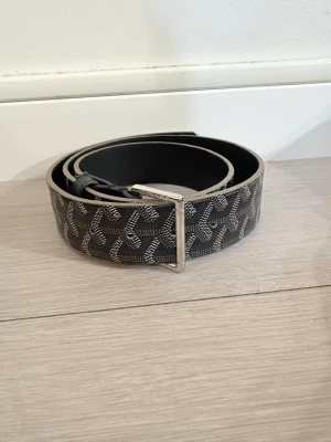 Svart mönstrad bälte med silverdetaljer - Snyggt svart bälte med ett grafiskt mönster i grått och silver. Bältet har en stilren silverfärgad metallspänne och är tillverkat i ett slitstarkt syntetmaterial med skinnliknande känsla. Perfekt för att lyfta din outfit med en cool detalj.