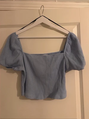 Ljusblå ribbad topp från H&M - Säljer en ljusblå ribbad topp från H&M i storlek L. Toppen har puffiga korta ärmar och fyrkantig halsringning, även bak. Materialet är mjukt och stretchigt, perfekt för vår och sommar. Enkel och stilren design som är lätt att matcha med jeans eller kjol. 
