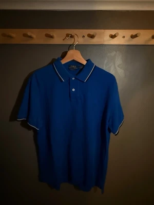 Blå Polo Ralph Lauren piké  - Snygg blå piké från Polo Ralph Lauren med klassisk pikékrage och vita detaljer på krage och ärmslut. Västen har två knappar framtill och är i en normal passform. Perfekt för dig som gillar stilrena och tidlösa plagg. Pris går att diskutera.