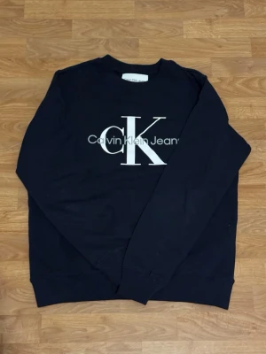 Svart sweatshirt från Calvin Klein - Snygg svart sweatshirt från Calvin Klein Jeans med stort CK-tryck i vitt på bröstet. Tröjan har rund hals, långa ärmar och ribbade muddar vid ärmslut och nederkant. Perfekt för en chill och stilren look.