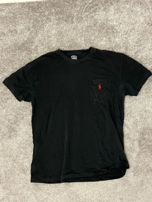 Svart t-shirt från Polo Ralph Lauren - Svart t-shirt från Polo Ralph Lauren med klassisk rund hals och korta ärmar. På bröstet finns en ficka med röd broderad Polo-logga. T-shirten är gjord i mjuk bomull och har en clean, stilren look som funkar till allt. Tröjan är använd så slitage finns 