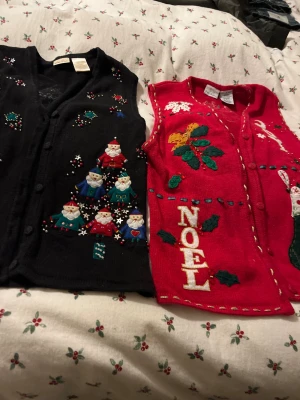 Juliga cardigan jackor med broderi - Två cardigan jackor, en svart och en röd, båda med julbroderier och pärlor. Den svarta har små tomtar i form av en julgran, och den röda har texten NOEL, mistel och julmotiv. Båda är ärmlösa och har knappar framtill. Perfekt för att maxa julstämningen!