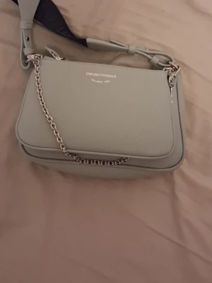 Blå axelväska från Emporio Armani - Snygg blå axelväska från Emporio Armani med stilren design och silverfärgad kedjedetalj. Väskan har ett brett, justerbart axelband i svart och grått, samt präglad logga på framsidan. Tillverkad i skinn med slät yta och praktisk dragkedja upptill. Säljer åt en vän, kvitto finns från köp för äkthets intygande, Pris går att diskuteras