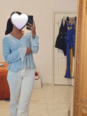 Ljusblå blus med volang och knappar - Supersöt ljusblå blus med v-ringning, knappar framtill och volangdetalj vid midjan. Ärmarna är långa och har en lätt utsvängd volangkant. Materialet är lätt och luftigt, perfekt för vår och sommar. Passar dig som gillar romantisk och feminin stil. Lite stor i storleken 