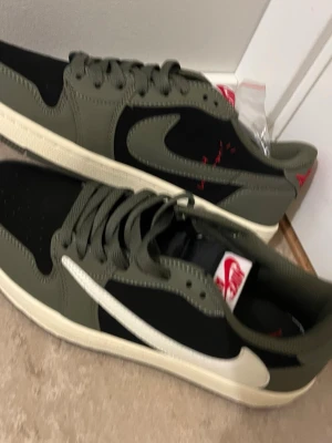 Nike Air Jordan 1 Low Travis Scott - Nike Air Jordan 1 Low Travis Scott i olivgrön och svart med vit swoosh och röda broderade detaljer på hälen. Skorna har snörning, platt sula och rund tå. Materialet är en mix av skinn och mocka, med klassisk Jordan-logga på baksidan.