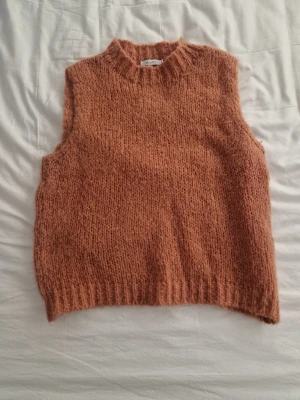 Stickad orange ärmlös tröja Noella - Säljer en mysig stickad ärmlös tröja från Noella i storlek M/L. Tröjan har en hög krage och är i en varm orange färg med grovstickad struktur. Perfekt att styla över en skjorta eller t-shirt för extra lager.